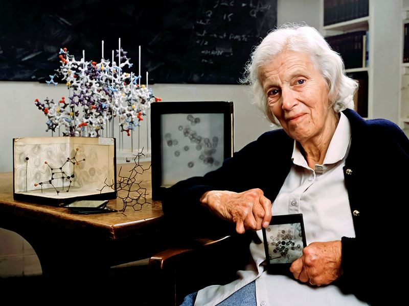Dorothy Hodgkin sits in front of some chemistry equipment and holds a chemistry-related award
