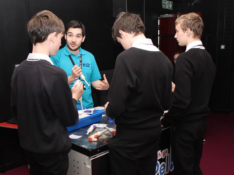 A volunteer holds a pipetting instrument and demonstrates its use to three boys in school uniform