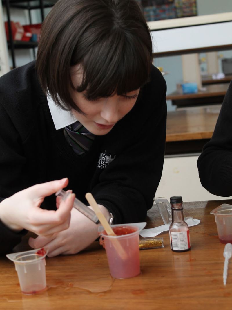 A girl in school uniform uses a syringe to injuect something into a beaker filled with a red substance.