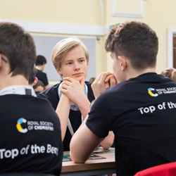 A student thinks about a question while two others, facing away from the camera wear Top of the Bench shirts, during the Top of the Bench final
