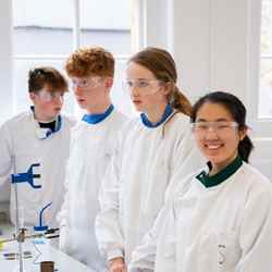 Four students, donning white coats and safety goggles, work away in the lab