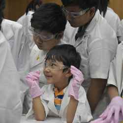 An excited young boy wearing safety goggles and gloves watches an experiment with other children