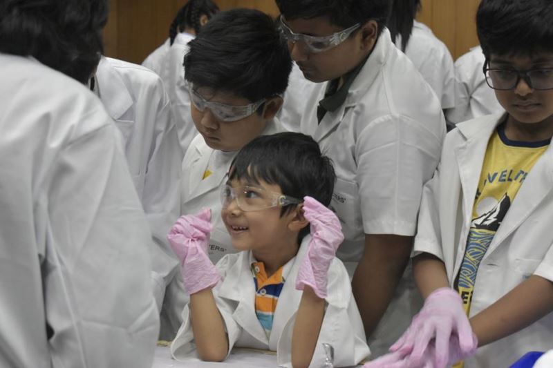 excited boy wearing safety goggles and gloves watches an experiment, surrounded by other children
