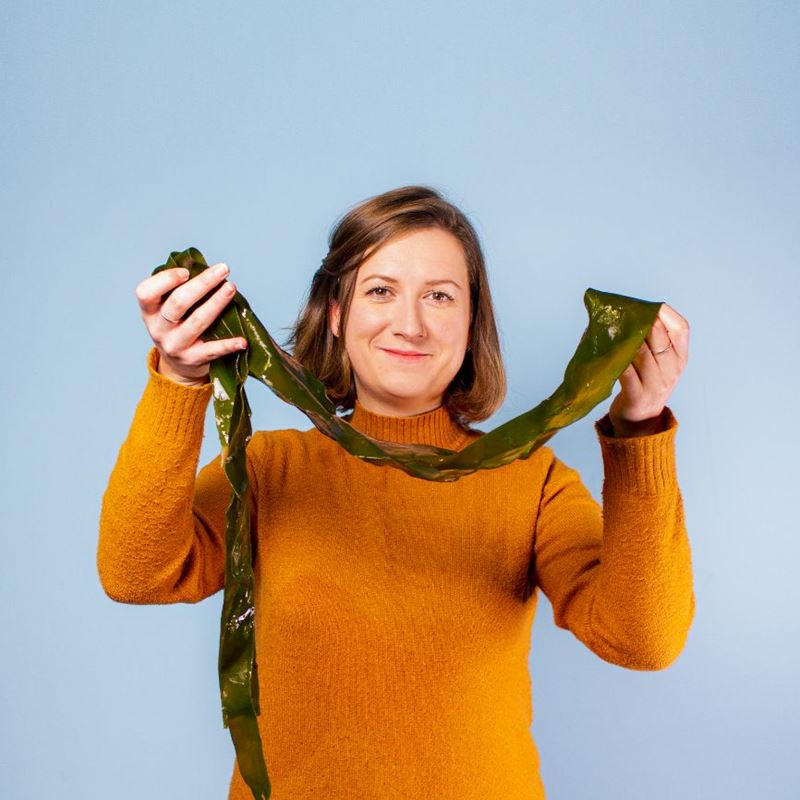 Louise Anderson, Research Director at Notpla, stands smiling and holding a long piece of seaweed