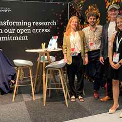 Three members of RSC staff, including Sara Bosshart (far right), stand next to a table with some stools and a banner saying "Transforming research with our open access commitment"