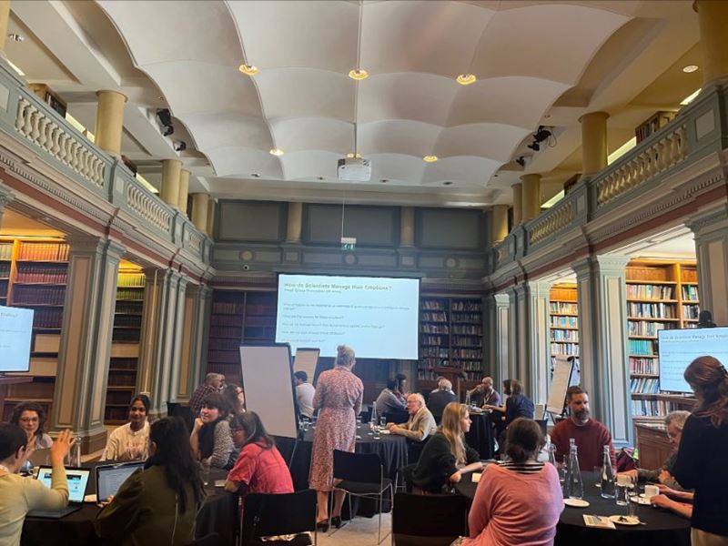 A landscape image of a grand room in Burlington House with books along the walls and event attendees sat around circular tables in discussion with a screen asking 'How do scientists manage their emotions?'