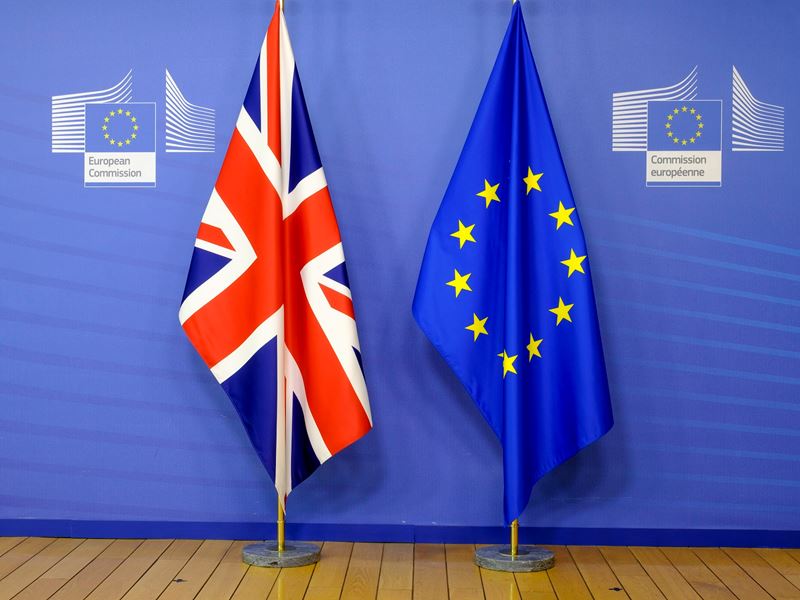 A Union Jack and an EU flag droop from flagpoles in front of a blue European Commission background and above a wooden floor