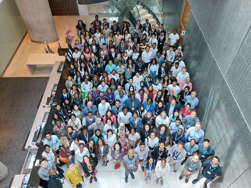 More than 130 students and graduates, plus numerous RSC and company representatives, stand together and look up towards the camera in a hotel lobby