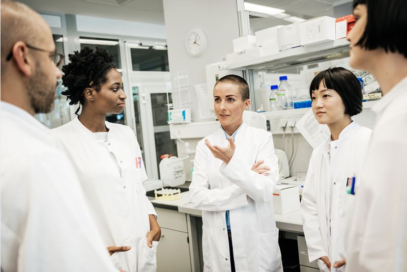A racially diverse group of five scientists wearing white coats stand and talk in a lab. A south Asian male, a black woman, a white woman, a south-east Asian female and a white woman are visible.
