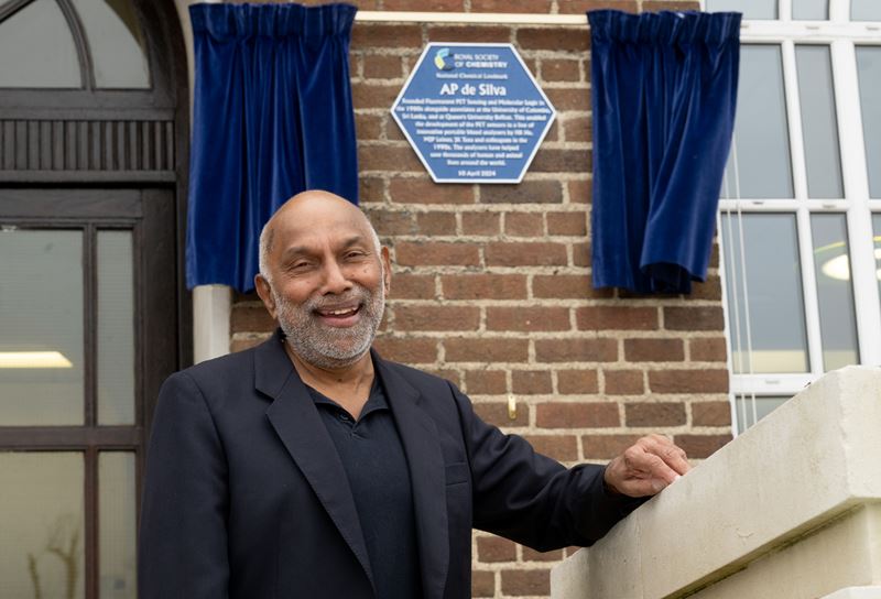 Prof AP de Silva smiles in front of his National Chemical Landmark plaque at Queen's University Belfast