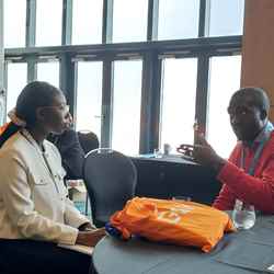 A female programme participant sits listening to a representative from GSK, while an orange GSK tote bag sits between them