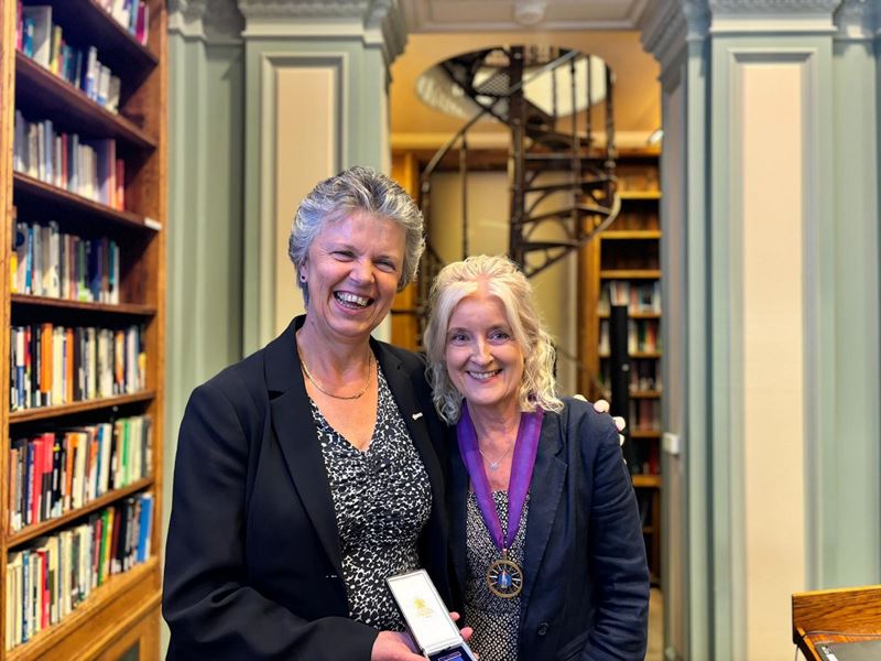 Professor Gill Reid smiles and hold a medal out while Dr Annette Doherty smiles at the camera