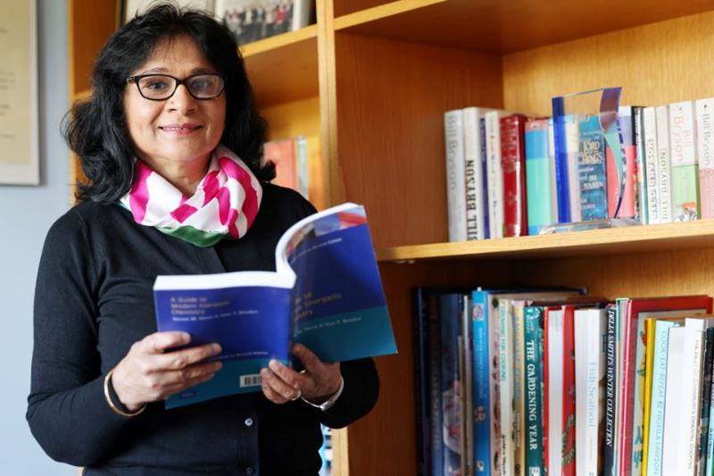 Professor Anju Massey-Brooker, industry associate at the Royal Society of Chemistry, smiles at the camera while holding a book open while stood in front of a bookcase
