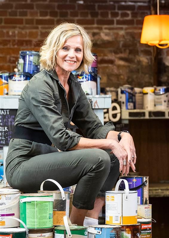 Linda Barker in green boiler suit sitting on stack of paint pots