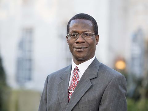 Professor Robert Mokaya smiles at the camera as he stands a slight angle, wears a grey suit, white shirt, red patterned tie and glasses